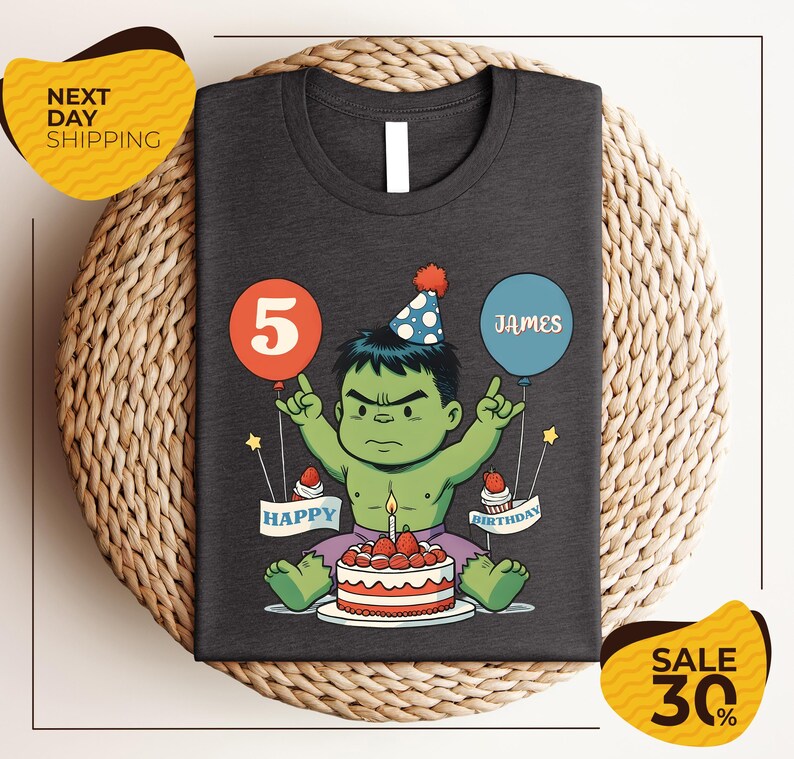 A t-shirt with a graphic design depicting a cartoon character wearing a party hat and holding a birthday cake, with balloons and the text "Happy Birthday James" around the character.