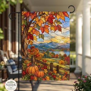 Puede incluir: Una bandera de jardín rectangular con una ilustración de un paisaje otoñal al estilo de vidrieras. La escena muestra un árbol con hojas naranjas y rojas, dos calabazas junto a una valla de madera, un lago y montañas lejanas al atardecer.