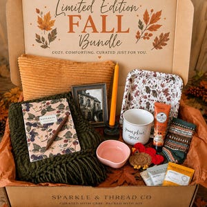 May include: A cardboard gift box labeled Limited Edition Fall Bundle containing a mustard corduroy pillow, a dark green knit throw, a notebook with a berry print, a brown pen, a framed house print, an orange taper candle, a white mug, a pink bowl, a crochet turkey, a patterned tray, a tube of lotion, and socks.