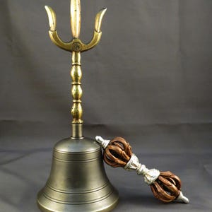 Puede incluir: Una campana de bronce con una parte superior en forma de tridente y un asa decorativa. Un vajra, u objeto ritual, de color cobre y plata descansa junto a la campana. La campana mide unos 15 cm de alto.