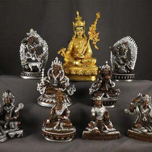 Könnte beinhalten: Eine Sammlung verzierter Statuen, die verschiedene Figuren darstellen, hauptsächlich in Gold-, Silber- und Bronzetönen. Die Statuen variieren in Größe und Design, einige weisen aufwendige Details und aufwendige Kopfbedeckungen auf. Die Figuren sind auf einer grauen Oberfläche vor dunklem Hintergrund angeordnet.