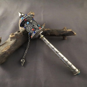 Puede incluir: Una rueda de oración plateada con intrincados filigranas y coloridos detalles de piedras preciosas. El cuerpo cilíndrico presenta patrones ornamentados y está adornado con piedras rojas, azules y turquesas. Una cadena con un dije decorativo cuelga de la rueda.