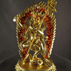 May include: A golden statue of a deity with multiple arms, adorned with intricate details. The figure holds a staff and is surrounded by a fiery, orange and gold halo. The statue is on a decorative base, showcasing a blend of artistry and religious symbolism.