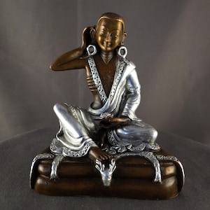 Può includere: Statua in bronzo e argento di una figura seduta con espressione serena. La figura indossa una veste argentata con dettagli intricati e grandi orecchini a cerchio. La statua è su una base marrone con accenti decorativi argentati.