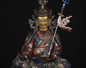 May include: A seated statue of a figure with a gold face and ornate headdress, wearing a colorful robe with silver accents. The figure holds a small bowl and a staff with a decorative top. The base is decorated with floral patterns.
