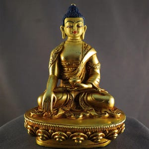 May include: A gold-coloured Buddha statue in a seated pose, with a blue topknot. The statue features detailed carvings on the robe and base, and holds a small bowl. The statue is set on a decorative base.