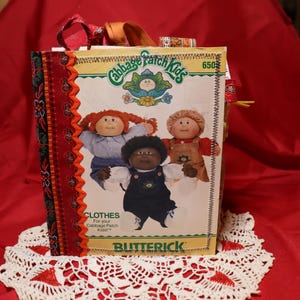 May include: A vintage Cabbage Patch Kids pattern book featuring three dolls on the cover. The book is on a white lace doily, with a red background. The book's spine is decorated with a floral pattern in red, orange, and green.