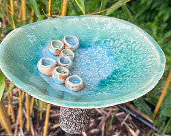 May include: A handcrafted ceramic bird bath with a turquoise glaze. The bowl features a textured surface and is adorned with small, round, ceramic elements. The bird bath is mounted on a decorative pedestal, set against a backdrop of bamboo.