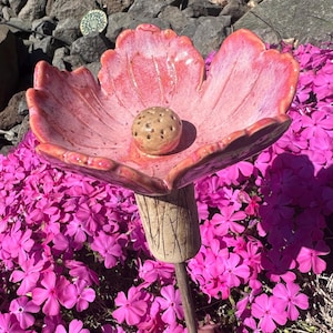Puede incluir: Una estaca de jardín de cerámica hecha a mano con un plato rosa en forma de pétalo y un tallo beige texturizado. El centro de la flor presenta una pequeña bola redonda perforada. La estaca se exhibe entre vibrantes flores rosas.