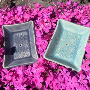 Könnte beinhalten: Zwei Keramik-Räucherstäbchenhalter, einer blau-grau und einer hellblau, jeweils mit einem Mandala-Design. Die Halter sind rechteckig mit gebogenen Kanten und einem zentralen Loch. Sie liegen auf einem Bett aus leuchtend rosa Blüten.