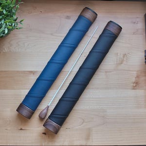 Könnte beinhalten: Zwei Dirigentenstäbe, einer blau und einer schwarz, mit Holzenden und Griffen. Zwischen ihnen liegt ein weißer Stab. Die Stäbe liegen auf einer hellen Holzoberfläche. Der blaue Stab hat einen Holzgriff und einen blauen Ledergriff.