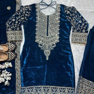 Könnte beinhalten: Ein teal-farbenes Salwar-Kameez-Set aus Samt mit aufwendiger silberner Stickerei. Die langärmlige Tunika hat detaillierte Blumenmuster auf den Ärmeln und am Ausschnitt, mit passenden Verzierungen am Saum. Das Set enthält einen Schal und eine Hose.