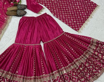 Può includere: Un completo salwar kameez indiano ricamato color magenta. Il set comprende una tunica, pantaloni a gamba larga e una sciarpa, tutti adornati con ricami dorati. L'outfit è esposto con sandali e fiori abbinati.