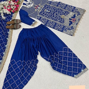 Op de afbeelding: Een koningsblauwe salwar kameez set met ingewikkelde zilveren borduursels. De set bevat een tuniek met lange mouwen, een loszittende broek en een bijpassende sjaal. De tuniek en broek hebben gedetailleerde patronen en versieringen, perfect voor speciale gelegenheden.
