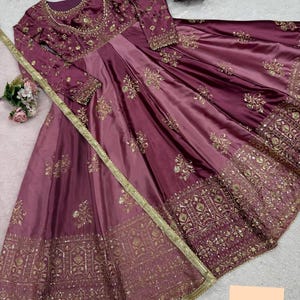 May include: A long, flowing, formal dress in shades of purple and mauve, adorned with intricate gold embroidery and sequins. The dress features long sleeves and a detailed bodice, with a wide skirt. A gold trim and a matching scarf complete the elegant design.
