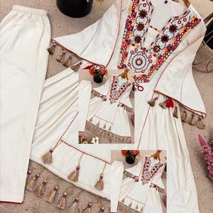 May include: A white embroidered tunic and matching trousers set. The tunic has a V-neck with colourful embroidery and decorative tassels along the hem and sleeves. The trousers are a solid white colour. The set is displayed on a neutral surface.