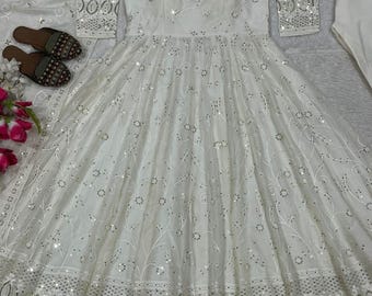 May include: A white, embellished dress with long sleeves and a flowing skirt. The dress features intricate embroidery and sparkling sequins. A matching white scarf and pants are also included. Brown sandals with decorative beading are placed near the dress.