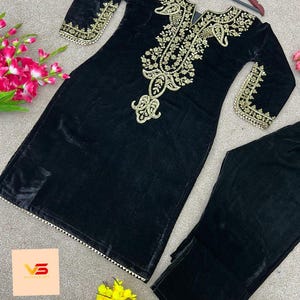 Puede incluir: Conjunto de túnica y pantalón de terciopelo negro. La túnica presenta bordados dorados alrededor del cuello, las mangas y el corpiño. El conjunto se exhibe sobre una superficie gris claro con detalles florales.