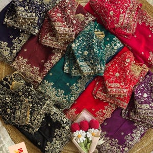 May include: A collection of elaborately embroidered blouses and matching fabric in shades of navy, burgundy, emerald green, and red. The garments display detailed gold threadwork and embellishments. A small bouquet of crocheted flowers is also visible.