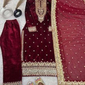 May include: A burgundy velvet salwar kameez set with gold embroidery and embellishments. The set includes a long-sleeved tunic, matching trousers, and a sheer dupatta. The outfit is displayed with a pair of colourful embroidered shoes.