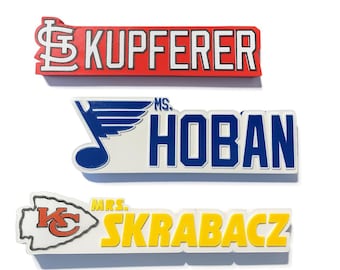 May include: Three name badges with team logos and names. The top badge is red with the name "KUPFERER" in white. The middle badge is white with a blue logo and the name "MS. HOBAN" in blue. The bottom badge is white with a yellow name "MRS. SKRABACZ".
