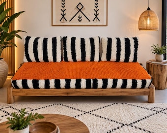 May include: A low-profile wooden daybed featuring a thick, textured orange seat cushion and three matching rectangular back cushions. The base and back cushions are decorated with a bold, vertical black and white striped pattern.