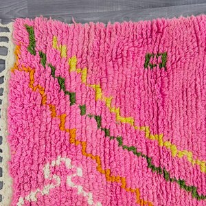 May include: A bright pink shag rug featuring a thick, high-pile texture and a series of diagonal, zigzagging lines in shades of green, yellow, and orange. The rug is finished with a cream-colored fringe along the left edge of the textile.