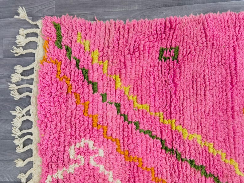 May include: A bright pink shag rug featuring a thick, high-pile texture and a series of diagonal, zigzagging lines in shades of green, yellow, and orange. The rug is finished with a cream-colored fringe along the left edge of the textile.