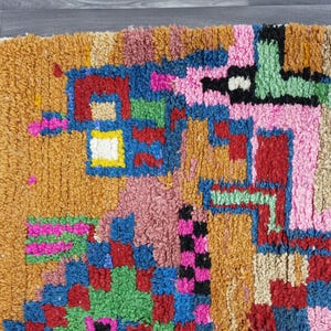 May include: Close-up of a colorful, textured rug with a geometric pattern. The rug features a mix of colors including orange, pink, blue, green, and red. The design is abstract and the rug has a soft, plush appearance.