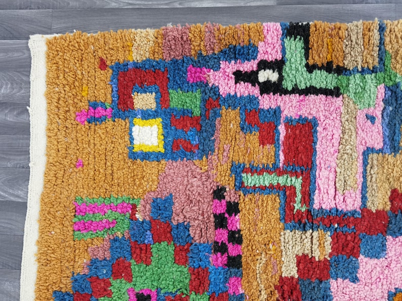 May include: Close-up of a colorful, textured rug with a geometric pattern. The rug features a mix of colors including orange, pink, blue, green, and red. The design is abstract and the rug has a soft, plush appearance.