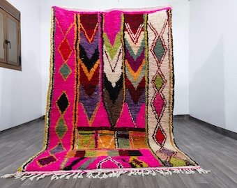 May include: A vibrant, hand-woven rug with a pink background and geometric patterns in various colors. The design includes diamonds, chevrons, and other shapes in shades of red, green, purple, and black. The rug has a fringed edge.