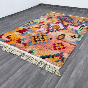 May include: A rectangular, hand-woven rug with a vibrant, geometric design. The rug features a mix of colors including orange, pink, blue, and green, with a cream-colored fringe along the edges. The rug's dimensions are not visible in the image.