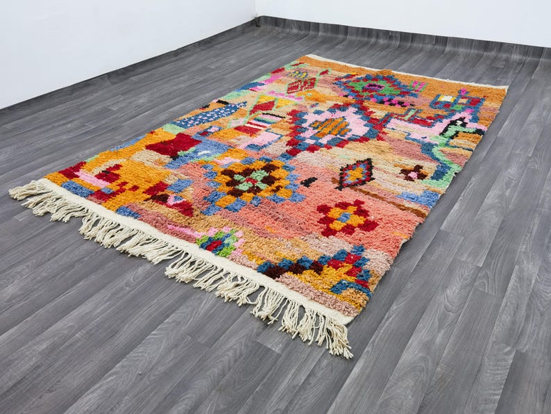 May include: A rectangular, hand-woven rug with a vibrant, geometric design. The rug features a mix of colors including orange, pink, blue, and green, with a cream-colored fringe along the edges. The rug's dimensions are not visible in the image.