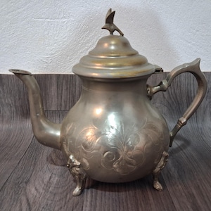 Puede incluir: Tetera antigua de color plateado con un adorno en forma de pájaro en la tapa. La tetera presenta grabados florales ornamentados y descansa sobre tres patas decorativas. El asa es curva y el pico es delgado.