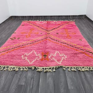May include: A rectangular area rug featuring a vibrant pink base with a geometric pattern in white, orange, and dark green. The design includes diamond shapes and cross motifs, finished with off-white knotted fringe along the two shorter edges.