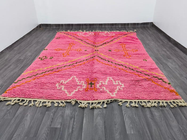 May include: A rectangular area rug featuring a vibrant pink base with a geometric pattern in white, orange, and dark green. The design includes diamond shapes and cross motifs, finished with off-white knotted fringe along the two shorter edges.