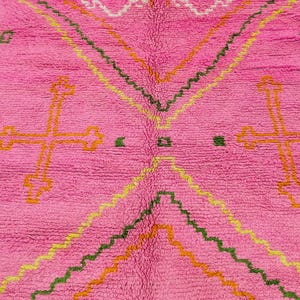 May include: A bright pink area rug featuring a high-pile texture and geometric patterns. The design includes large orange cross-shaped motifs, zigzagging lines in yellow and green, and small green square accents scattered across the surface.