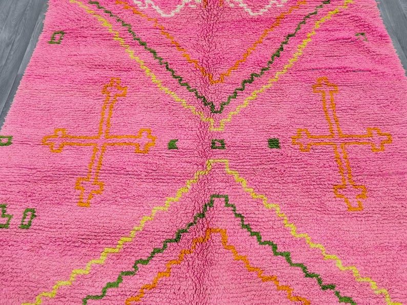 May include: A bright pink area rug featuring a high-pile texture and geometric patterns. The design includes large orange cross-shaped motifs, zigzagging lines in yellow and green, and small green square accents scattered across the surface.