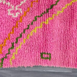 May include: A bright pink shag rug featuring a textured pile and off-white fringe along the edge. The surface is decorated with diagonal zigzag lines in orange, green, and yellow, along with small geometric shapes in white and orange.