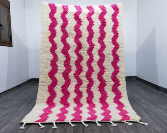 May include: A rectangular, cream-colored rug with a bold, zig-zag pattern in vibrant fuchsia. The rug has a thick, textured pile and white tassels along the bottom edge. The rug is displayed on a dark wood floor.