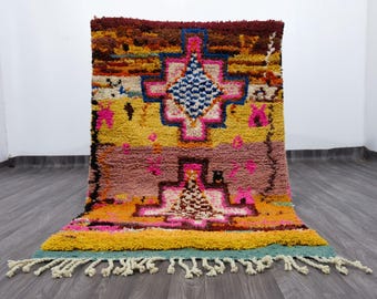 Puede incluir: Una colorida alfombra tejida a mano con un diseño geométrico. La alfombra presenta una mezcla de colores vibrantes, incluyendo amarillo, rosa, azul y marrón. Tiene una superficie texturizada y un fleco en un borde. Esta alfombra es adecuada para la decoración del hogar.