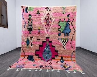 May include: A pink Moroccan rug with geometric designs in black, blue, green, and white. The rug has a textured appearance with a fringe along the bottom edge. The rug is displayed against a white wall.