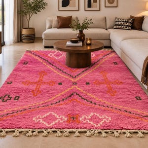 May include: A rectangular area rug featuring a vibrant pink base with a geometric pattern in white, orange, and dark green. The design includes large diamond shapes and cross motifs, finished with a cream-colored fringe along the short edges.
