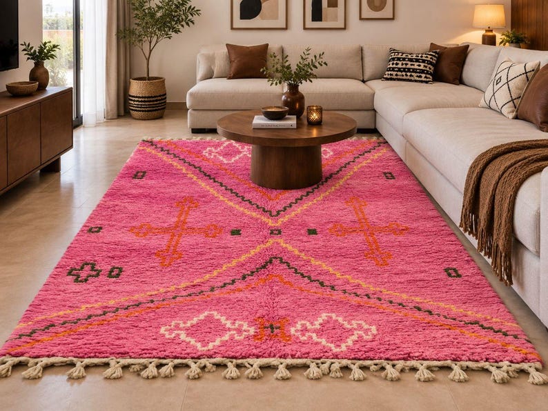 May include: A rectangular area rug featuring a vibrant pink base with a geometric pattern in white, orange, and dark green. The design includes large diamond shapes and cross motifs, finished with a cream-colored fringe along the short edges.