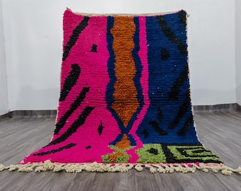May include: A vibrant, hand-woven rug with a bold geometric design. The rug features a striking combination of hot pink, navy blue, and burnt orange, accented with black and green shapes. The rug has a fringed edge and is displayed on a wooden floor.