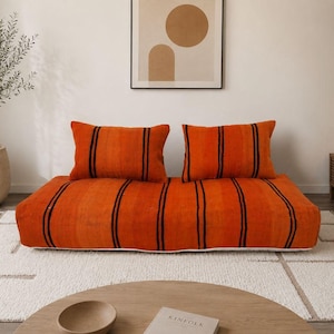 May include: A low-profile floor sofa featuring a vibrant orange fabric with vertical black stripes. The piece includes two matching rectangular back cushions and a wide, rectangular seat base, providing a simple, functional seating arrangement.