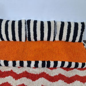 May include: A floor cushion with an orange centre and black and white striped border. It has three matching striped pillows. The cushion is on a rug with a red and white zig-zag pattern. The cushion measures approximately 100 cm.