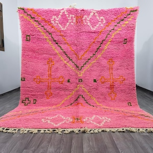 May include: A large, rectangular, bright pink area rug featuring a high-pile texture and a hand-knotted appearance. The rug displays geometric patterns in white, orange, and dark green, including cross-like motifs and diamond shapes, with a fringe edge.