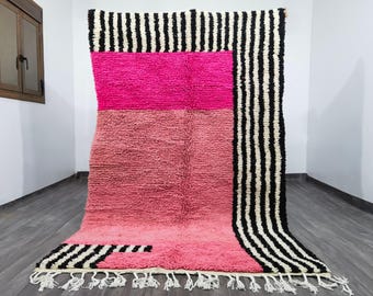 May include: A large, rectangular rug with a vibrant color scheme. The rug features a bold combination of pink and black and white stripes. The rug has a textured surface and a fringe along the bottom edge. The rug is made of a soft material.