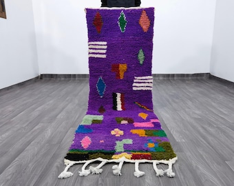 May include: A vibrant purple Moroccan rug with geometric designs. The rug features colourful diamond, rectangular, and floral patterns in shades of red, green, blue, and yellow. White tassels adorn the bottom edge. This handmade rug is a decorative floor covering.